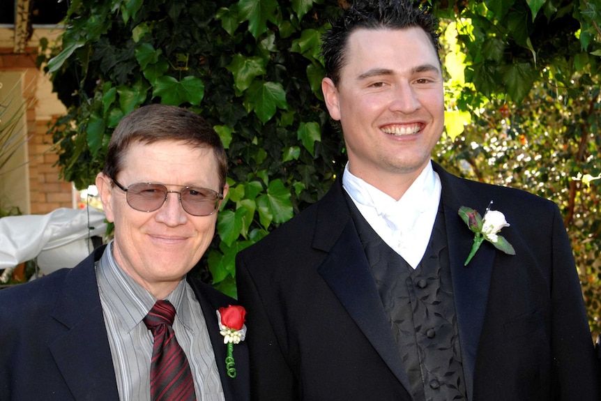 Two men, one in glasses, wearing suits and flower lapels, at a wedding.
