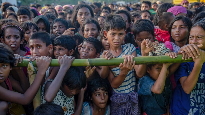 A crowd of Rohingya Muslim children, including a few in the front row carrying a bamboo stick.