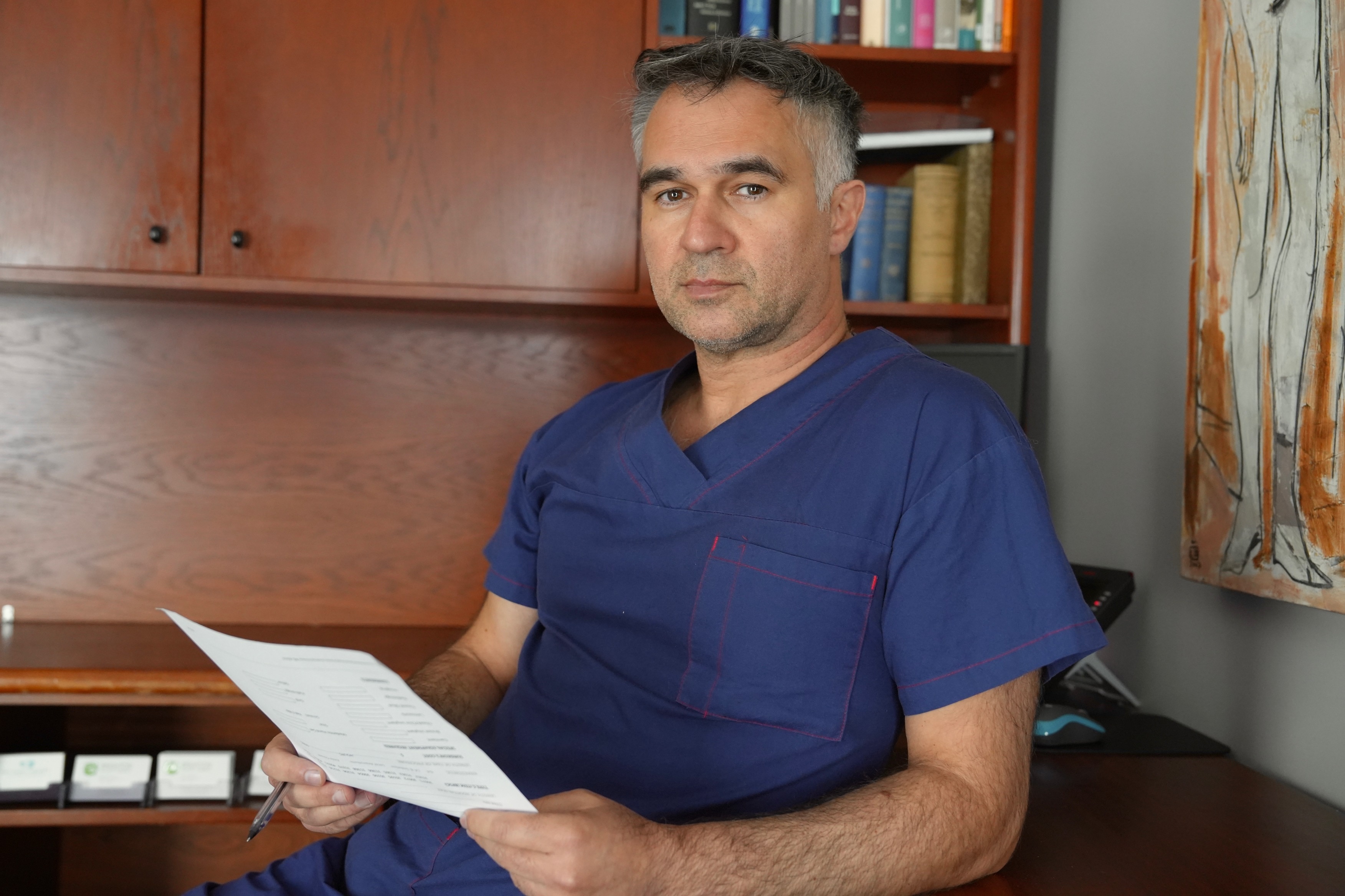 Uma foto mostrando um homem com uma bata de hospital azul olhando para a câmera.