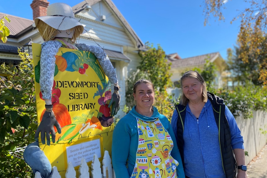 Two women standing next to a colourful sign reading Devonport seed library in front of a house.