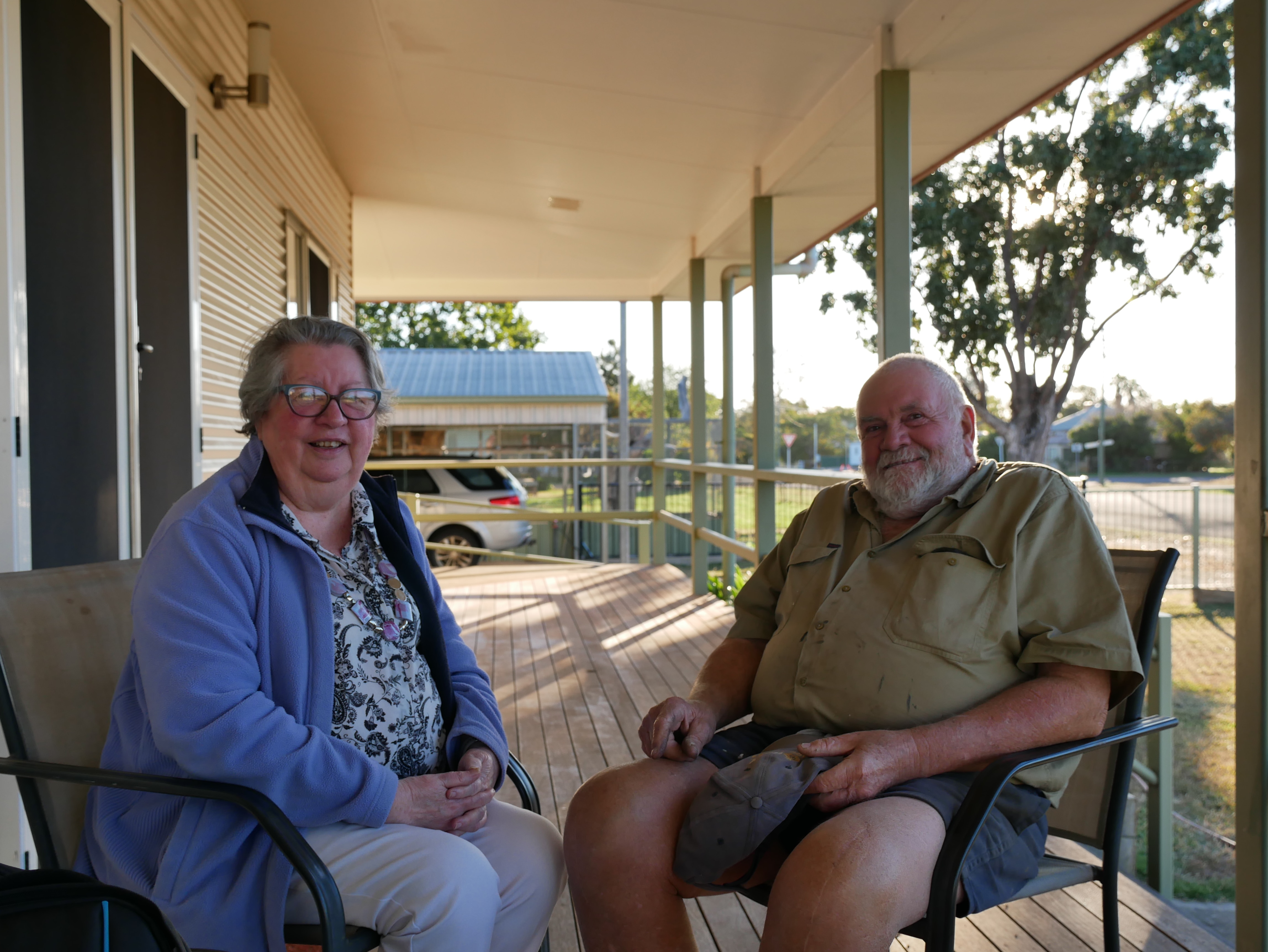 A man and a woman are sitting in chairs on the front porch of a house on a sunny day.