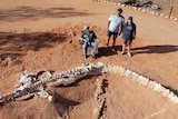Dino Dick sits in a chair next to the unearthed fossil, Tom and Sharron Hurley stand beside him.