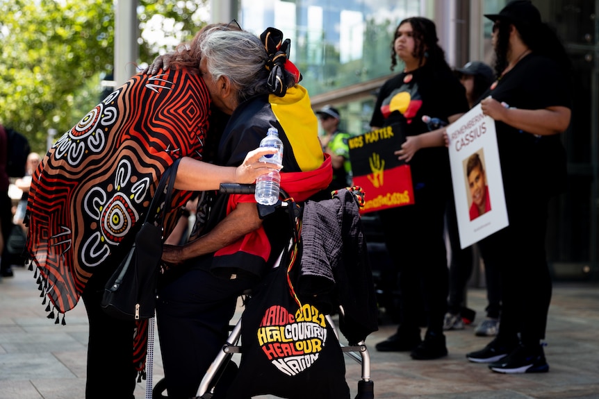 A woman in a wheel chair hugs another woman emotionally with people behind them holding placards.