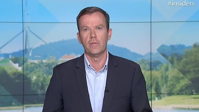 Wearing a suit jacket, Dan Tehan appears on television sitting in front of a mountain background