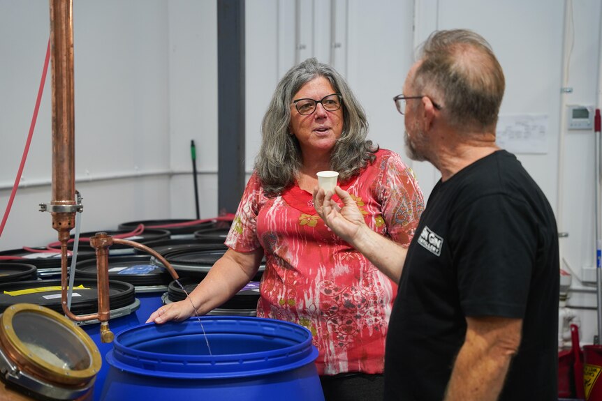 A middle-aged woman with grey hair and a red top stands over a blue barrel talking to a man in a black shirt.