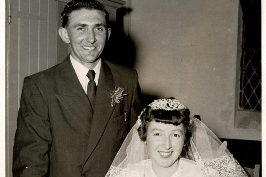 A black and white photo of a man in a suit standing next to a woman wearing a bridal gown sitting in front of a desk  