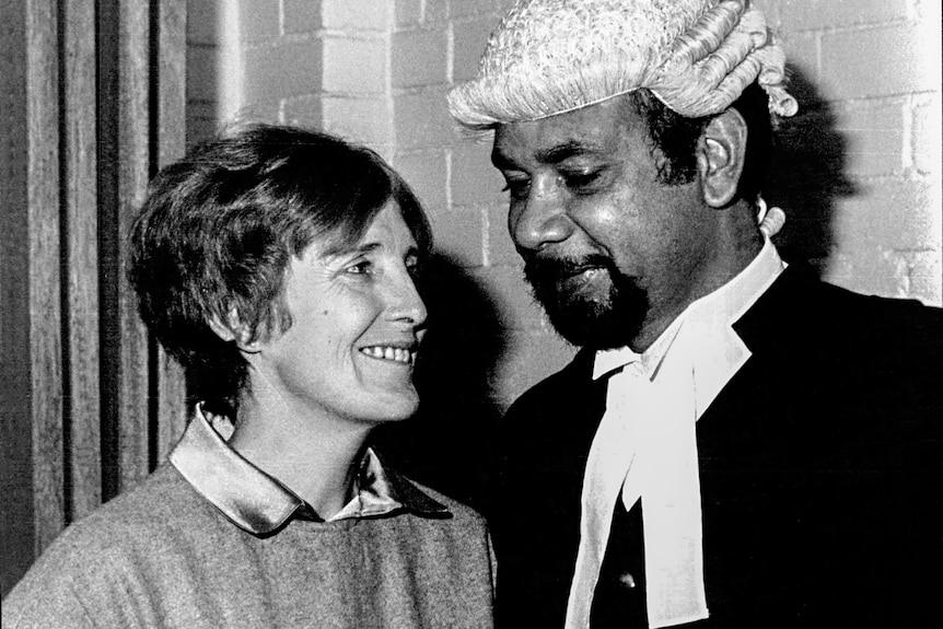A woman with short hair smiling at a man standing next to her in a judge's wig and cloak.