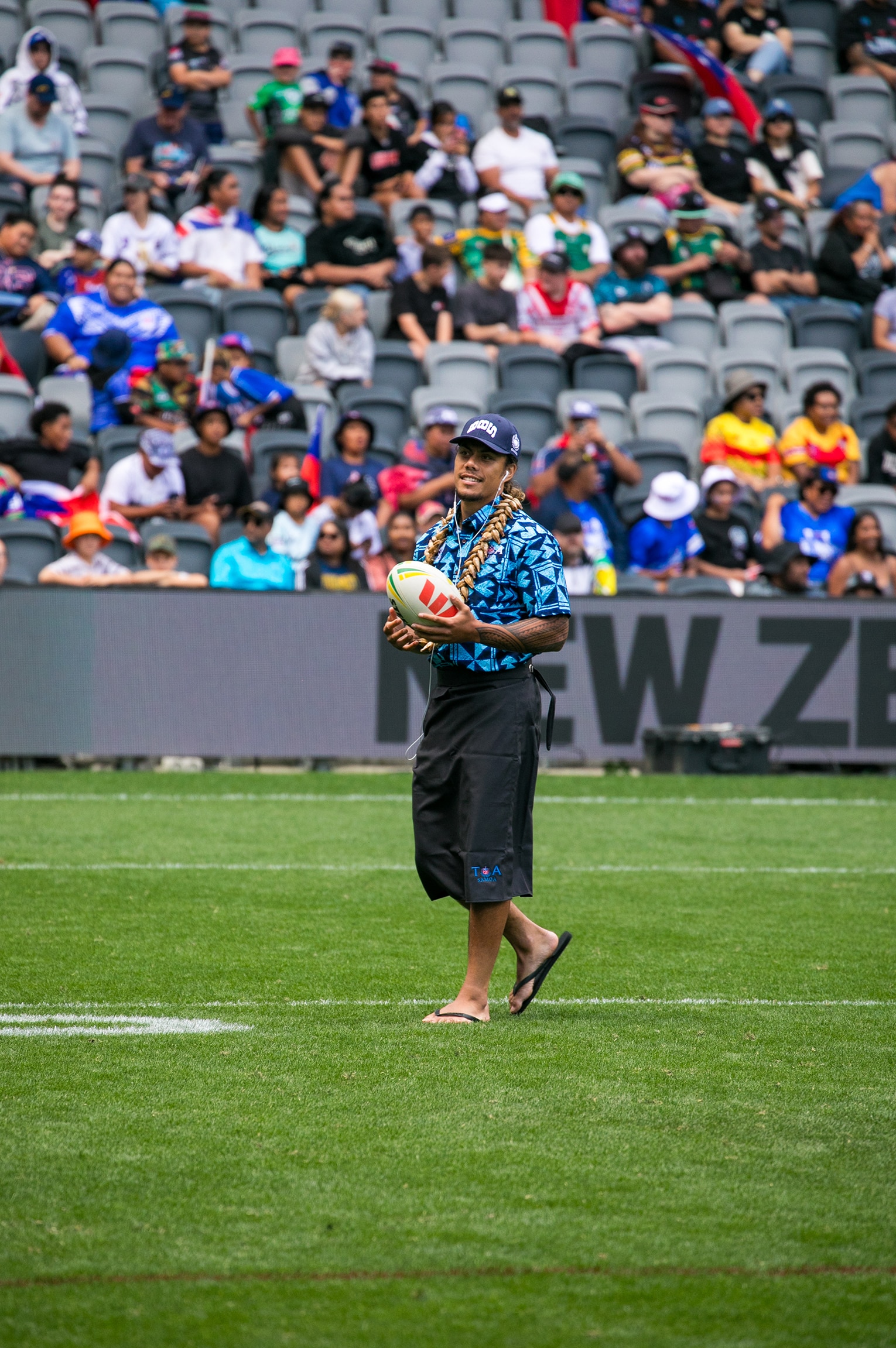 A man wearing a lavalava walks on a sporting field, holding a football.