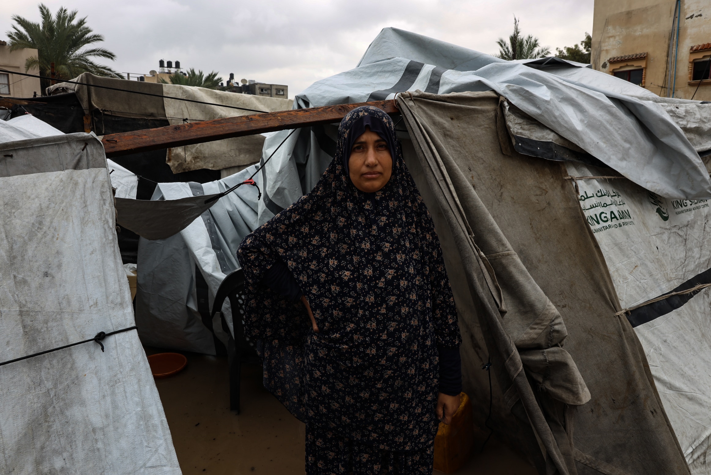 A woman wearing a hijab stands in front of makeshift tents with her hand on her waist. Muddy water is covering the ground.