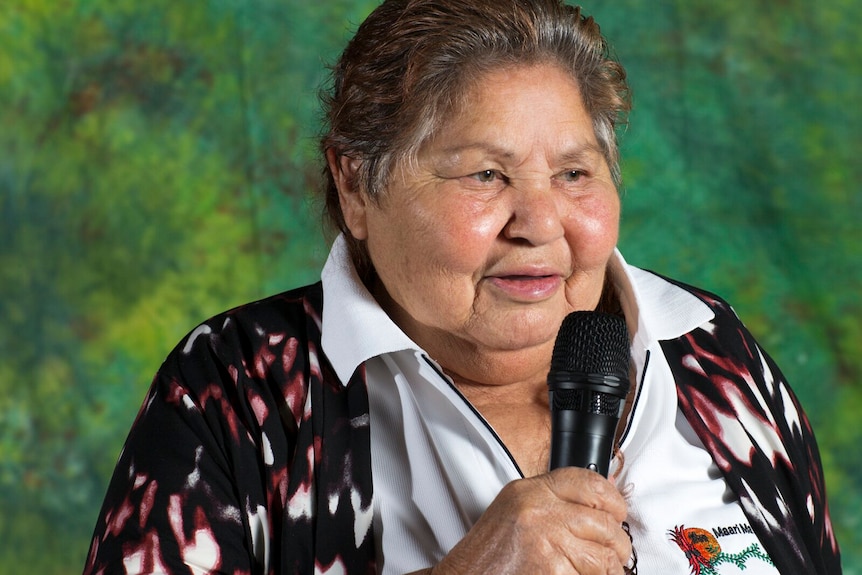 an older indigenous woman with short hair wearing a white shirt and colourful vest holding a microphone