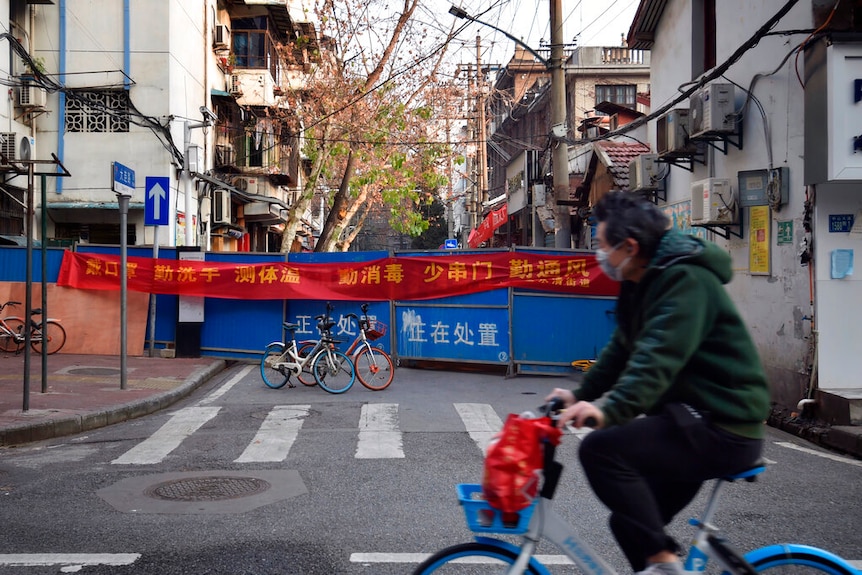 A man wearing a facemask cycles past an intersection with a bright blue barricaded fence blocking off a residential area.