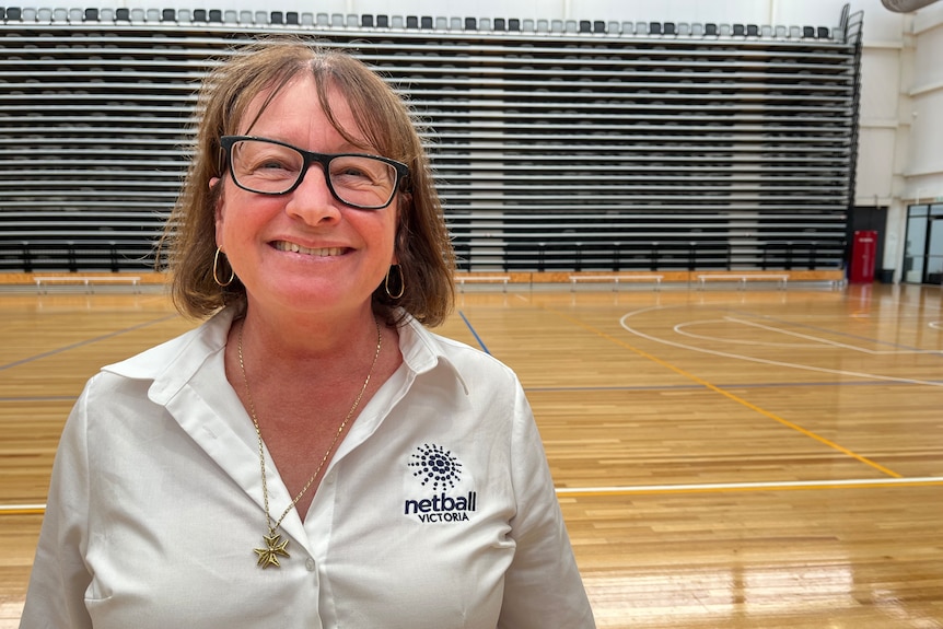 Judi has short brown hair and black glasses she stands on an indoor netball court and is wearing a maltese cross necklace