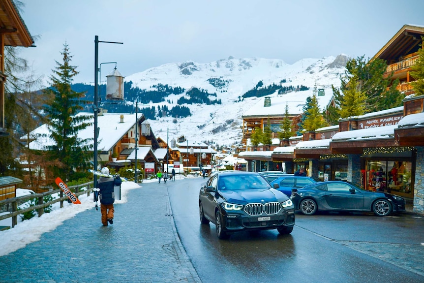 A snowboarder walks down a street in a snowy village at the base of a mountain