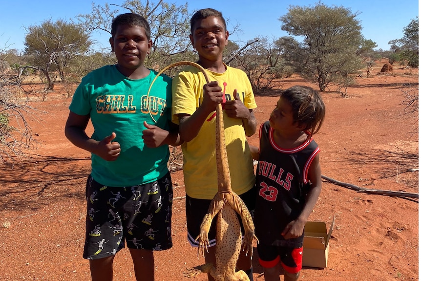 Three young boys stand proudly in a red desert, hoisting up a large yellow lizard.