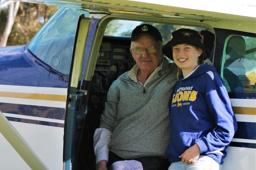 A smiling teenage girl stands close to her grandfather in the open door of a light aircraft
