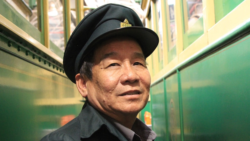 A smiling man in an old tram conductors uniform stands between two trams.