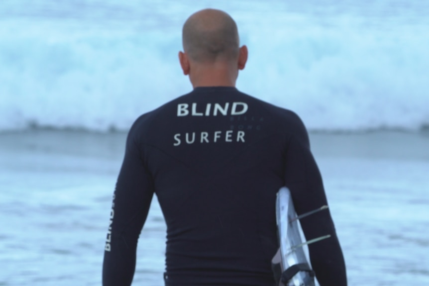 A man in a black wetsuit with white words 'Blind Surfer' walking towards waves with a board under his arm