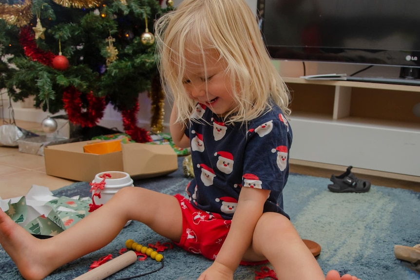 A smiling toddler wearing Christmas pyjamas opens presents.