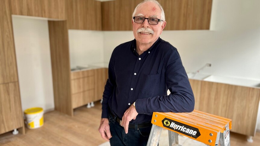 A man in his seventies leans against a ladder in a half renovated kitchen. 
