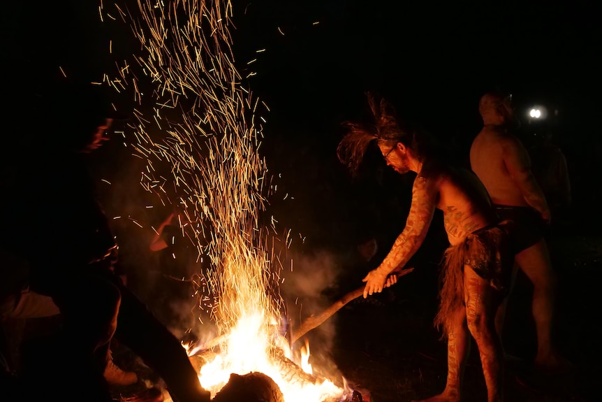 A man in traditional paint stokes a fire.