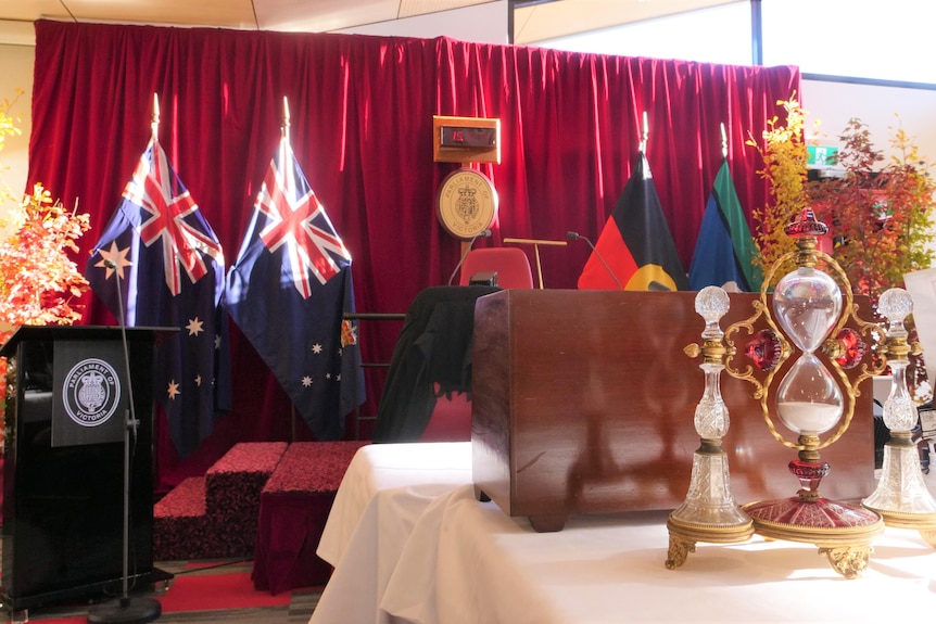 Four flags are behind a podium with a old fashion timer sitting on a table in foreground. 