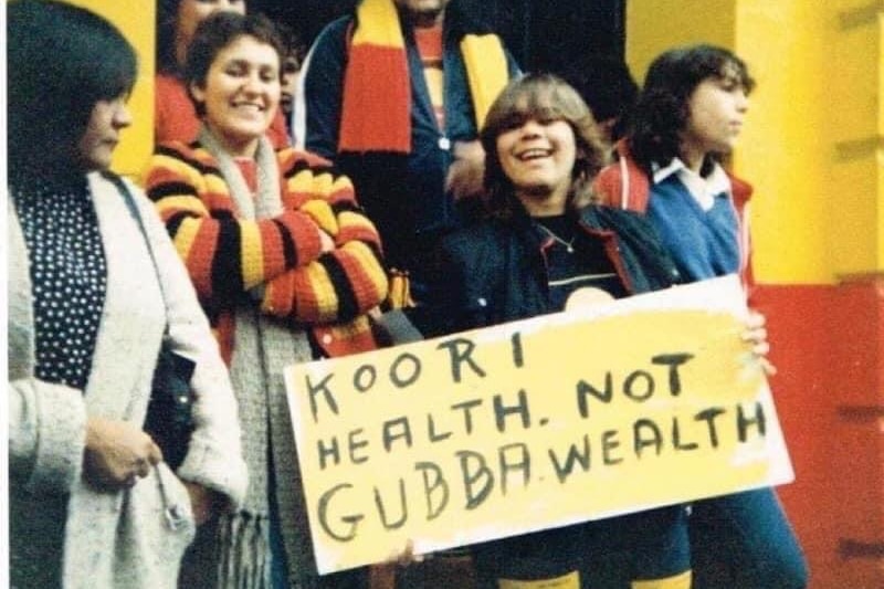 Men and woman stand outside a black, yellow and red coloured building. A sign reads 'KOORI HEALTH NOT GUBBAWEALTH'.