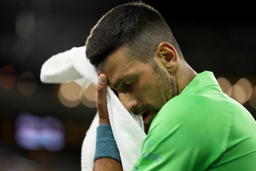 A tired-looking Tennis star Novak Djokovic sits with eyes closed as he holds a towel to his face during a match.