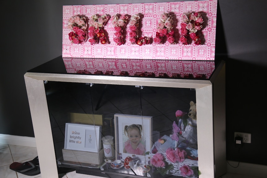 A pink sign with Skylar spelled out in pink and white flowers sits on top of a glass cabinet with a photograph of a little girl
