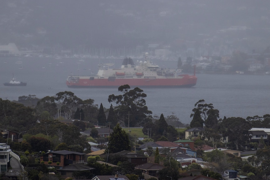 A red ship makes its way down a river with houses visible on either bank in rainy weather.