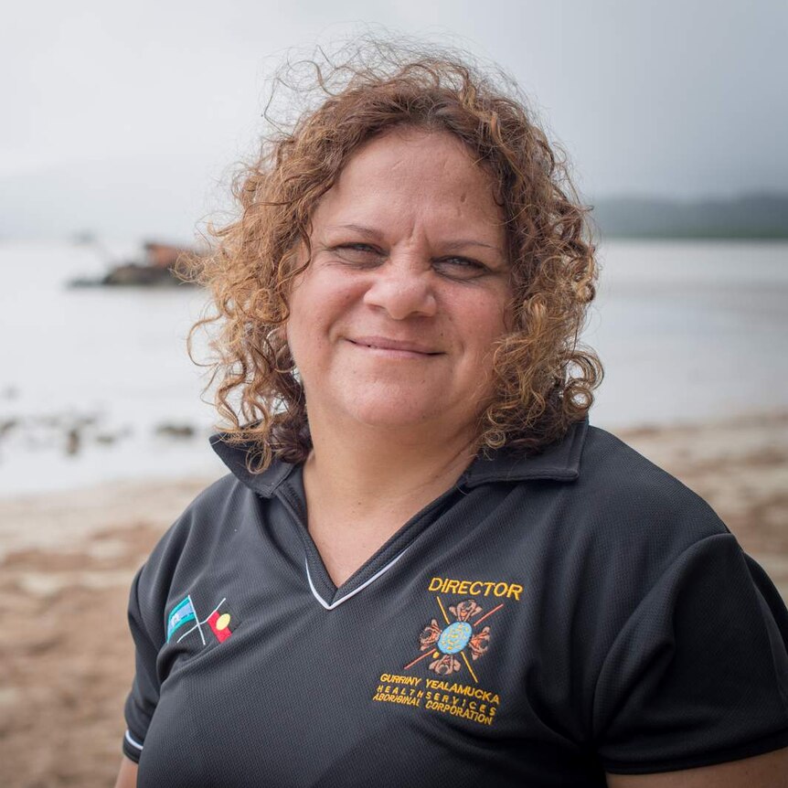 An Indigenous woman with shoulder-length curly hair, wearing a navy blue shirt, standing on a beach.