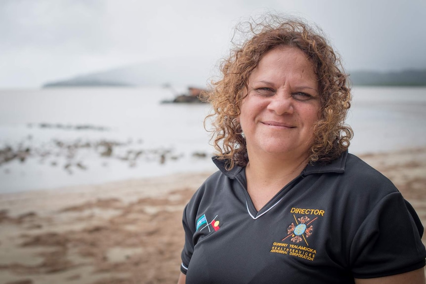 An Indigenous woman with shoulder-length curly hair, wearing a navy blue shirt, standing on a beach.