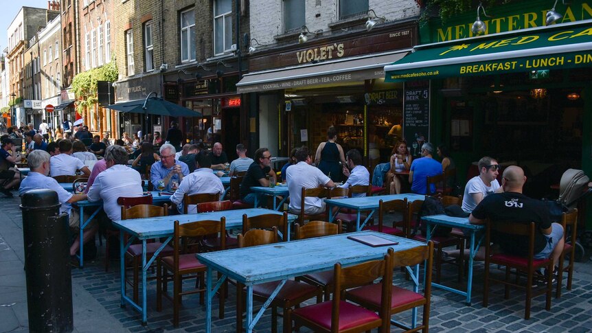 People sit at tables outside restaurants in a shady street.