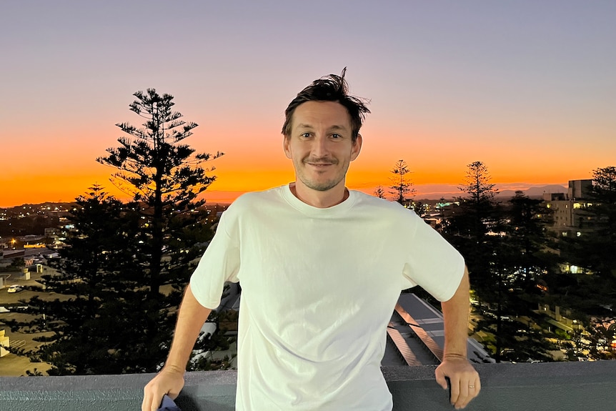Ένας άνδρας στα 30 του στέκεται σε ένα μπαλκόνι με μια περιφερειακή πόλη και το ηλιοβασίλεμα πίσω του.