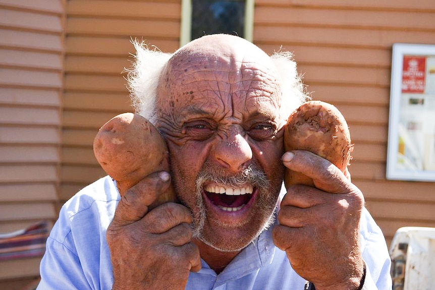 A smiling older man holding a potato in each hand holding them next to his head to depict tips for buying and cooking potatoes.