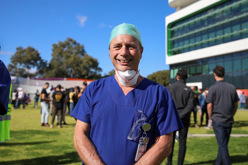 A man in health staff uniform with a mask on stands in a crowd outside a hospital.