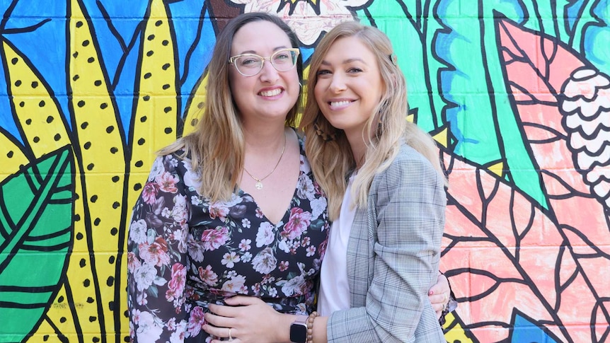 Profile picture of two young women hugging in front of brightly coloured wall.