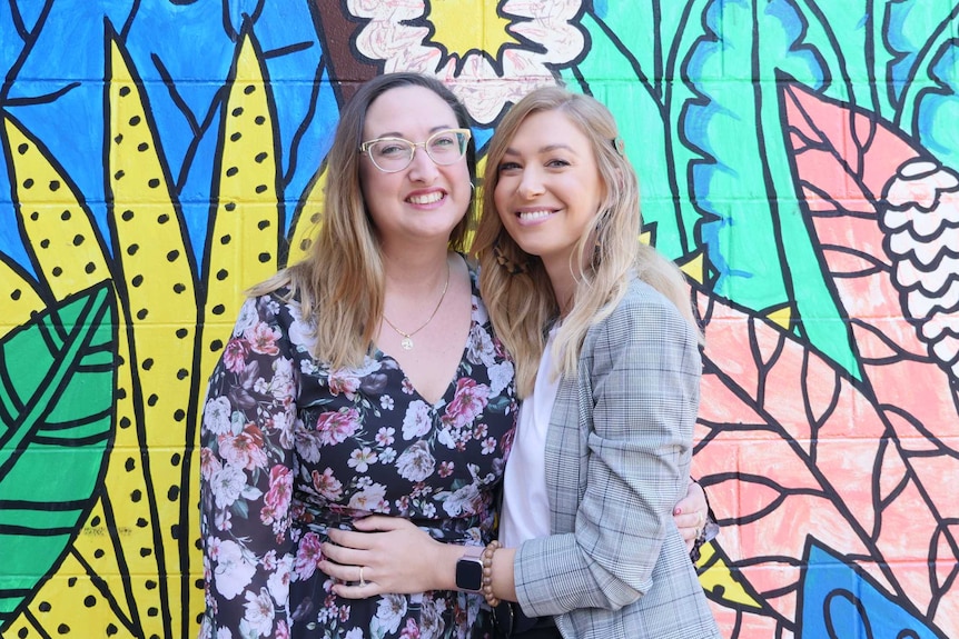 Profile picture of two young women hugging in front of brightly coloured wall.