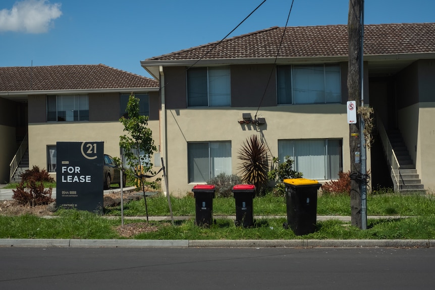 An exterior view of two houses close to each other with a 'For Lease' sign and wheelie bins out the front.