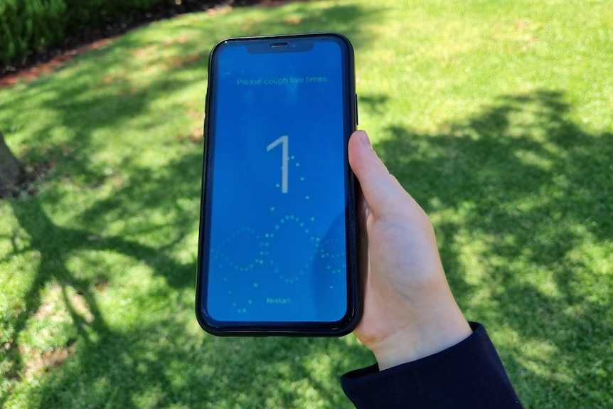 A smartphone being held in a child's hand with a blue cough screening application open, green lawn in background