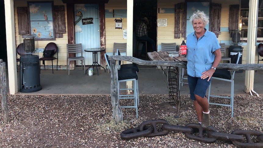 Woman wearing shorts and shirt holding a beer and smiling outside an old tin roofed pub with sign saying Hebel Hotel. 