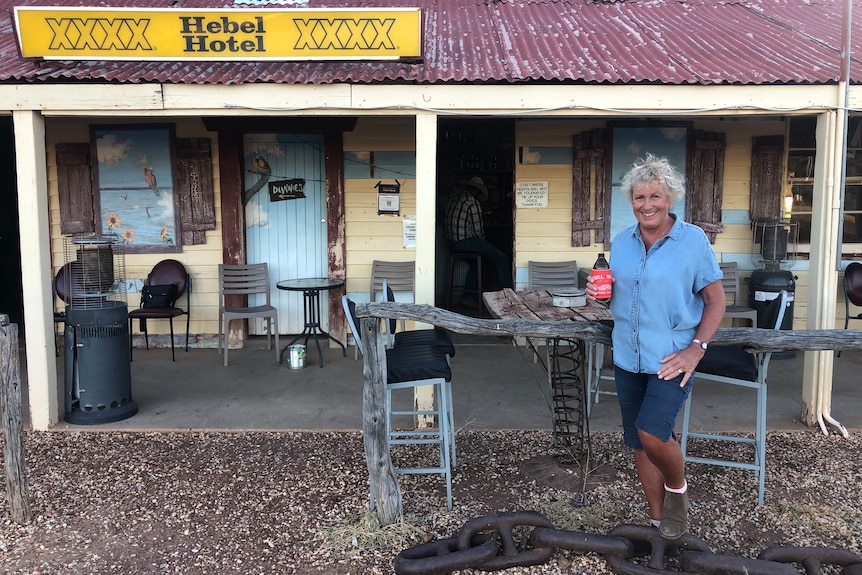 Woman wearing shorts and shirt holding a beer and smiling outside an old tin roofed pub with sign saying Hebel Hotel. 