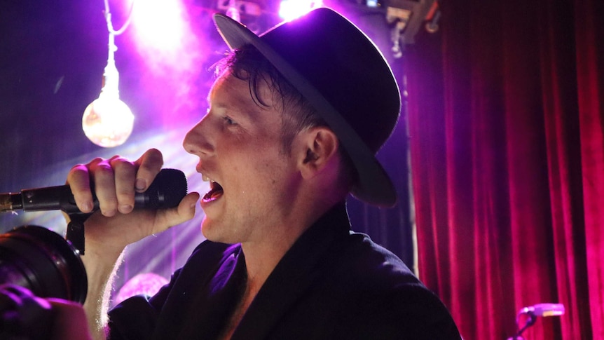 A man wearing a fedora hot singing with a microphone on stage, lights in the background