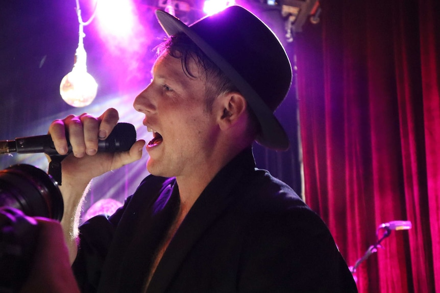 A man wearing a fedora hot singing with a microphone on stage, lights in the background