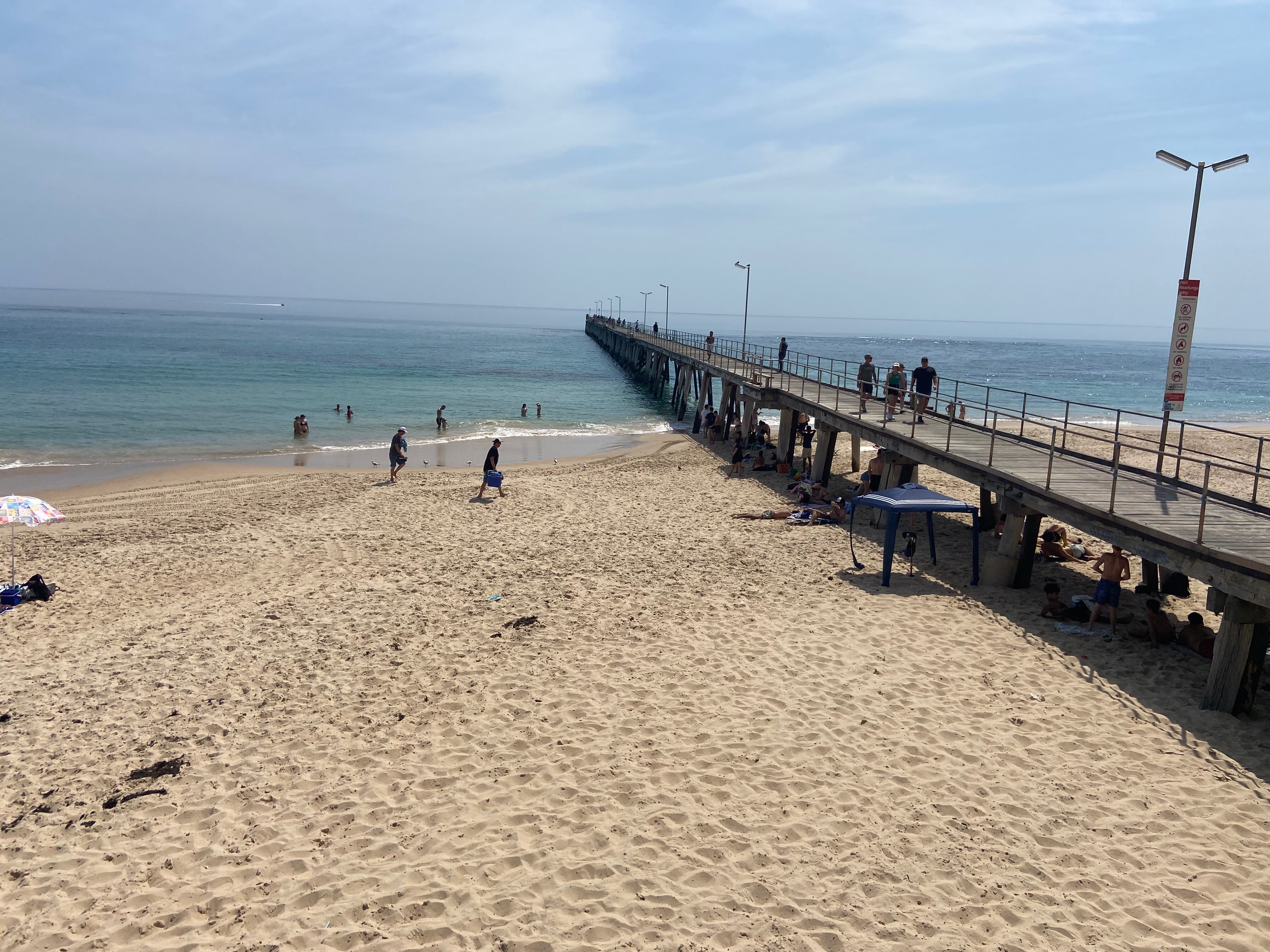 A sunny day at the beach, with people in the water and sitting under the jetty. 