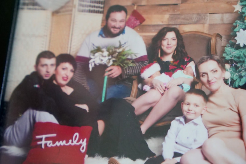 a family photo showing five adults and two kids sitting together smiling at the camera. A brown-haired woman is holding twins