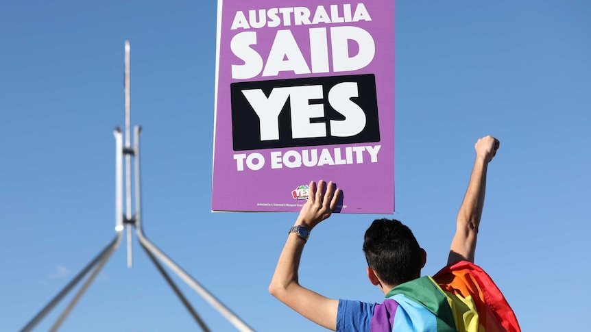 A person draped in a rainbow flag holds up a purple sign in front of the Parliament that says "Australia said yes to equality"