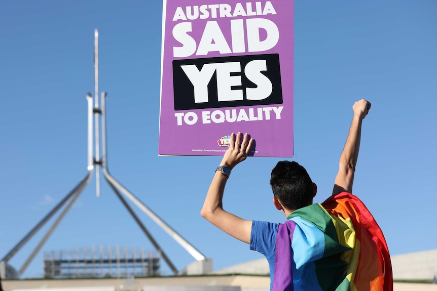 A person draped in a rainbow flag holds up a purple sign in front of the Parliament that says "Australia said yes to equality"