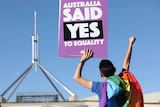 A person draped in a rainbow flag holds up a purple sign in front of the Parliament that says "Australia said yes to equality"