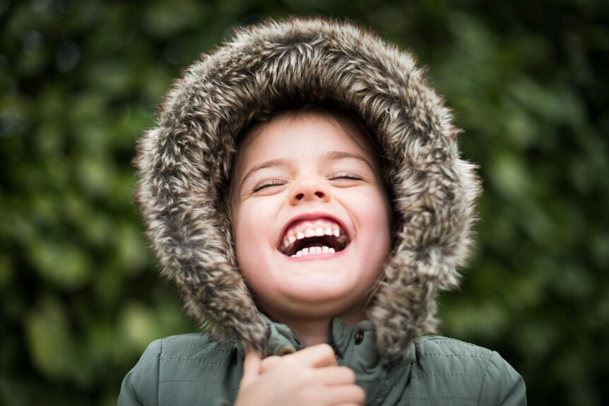 A kid throws his head back in laughter, he is wearing a dark green hoodie with a brown fur trim. 