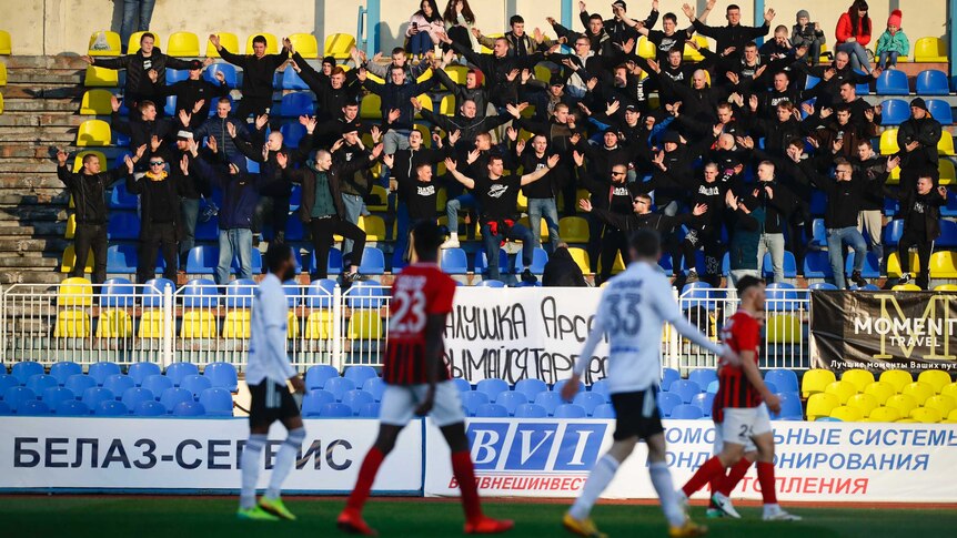 Fans stand with their arms outstretched in a grandstand with blue and yellow seats